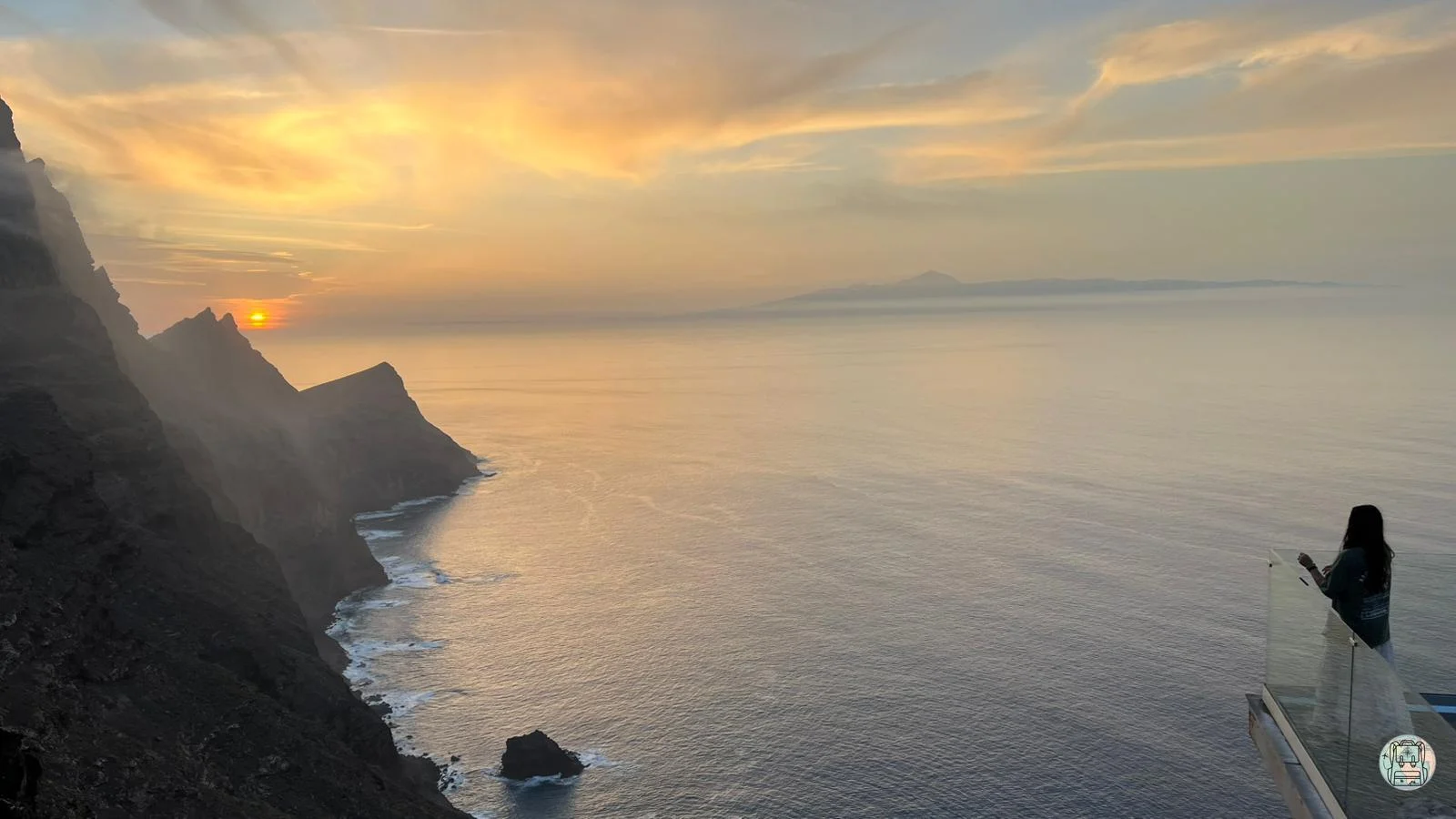 Vista panorámica de la costa escarpada de Gran Canaria al atardecer, con una persona en un mirador de cristal mirando el sol poniéndose sobre los acantilados y el Teide visible al fondo.