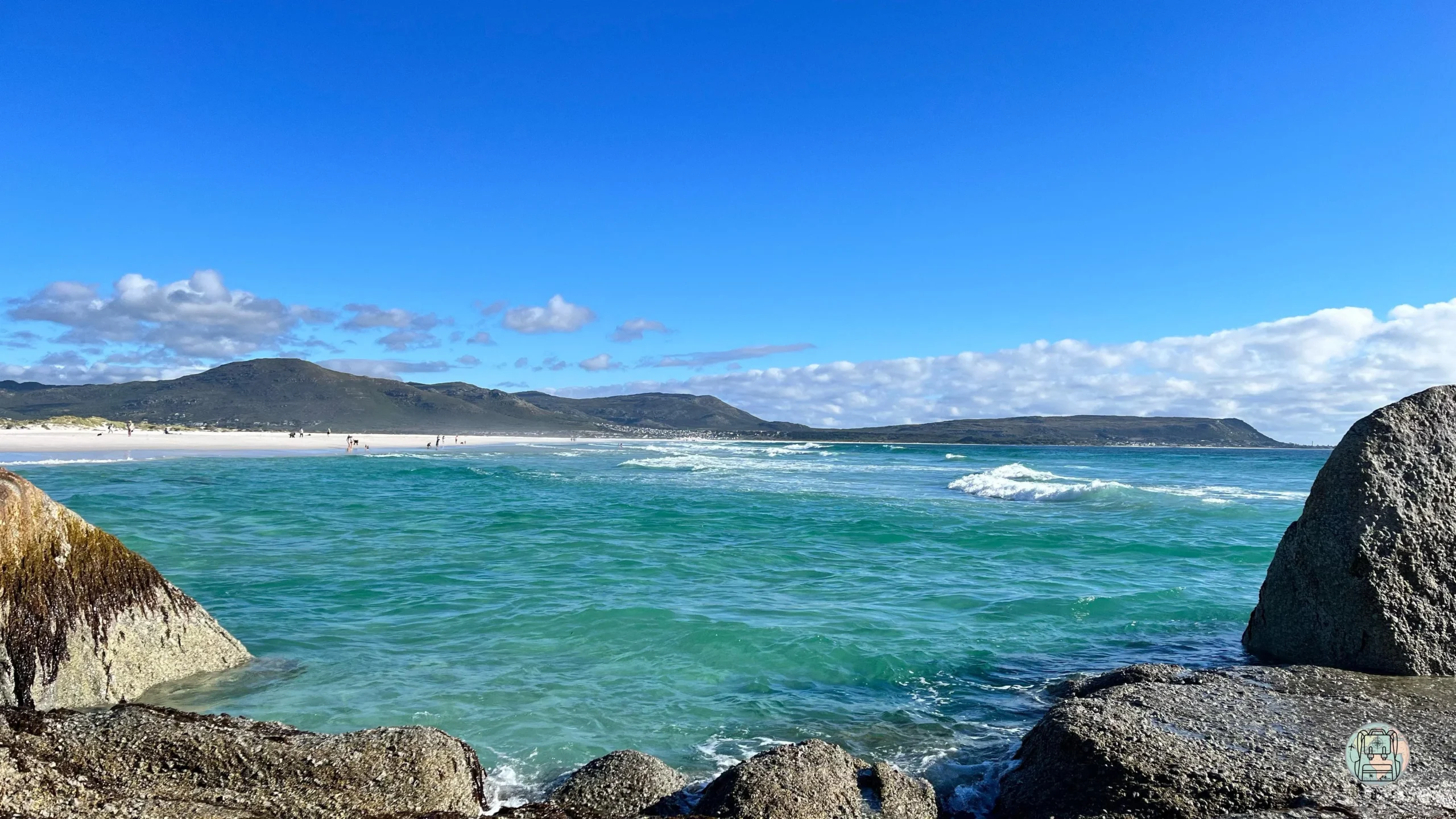 Aguas turquesas entre rocas de granito con vista a la extensa playa de Noordhoek y colinas al fondo